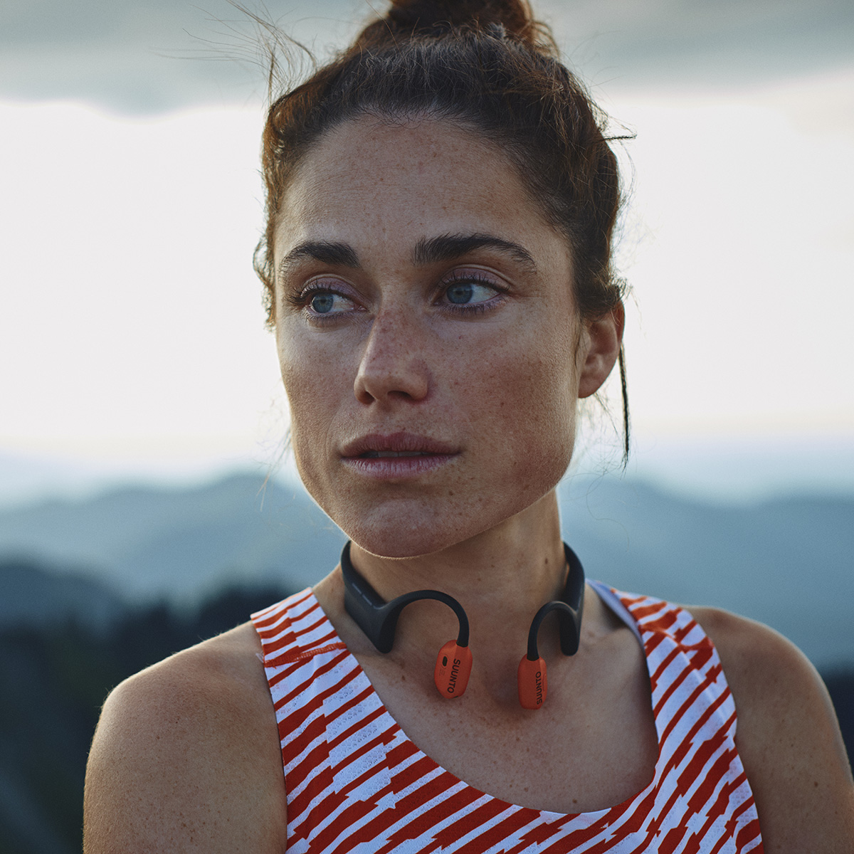 Frau mit Dutt und Sommersprossen trägt ein Headset. Rot-weiß gestreiftes Oberteil. Berge im Hintergrund. Der Himmel ist bedeckt.