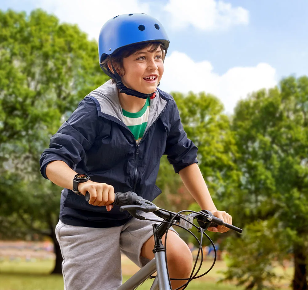 Chłopiec w niebieskim kasku jeździ na rowerze w parku. Ma kurtkę i szorty. W tle drzewa na tle błękitnego nieba.