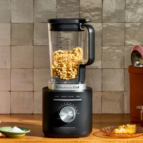 Egy KitchenAid turmixgép tele földimogyoróval egy fa felületen áll. Méztányér és sóstál közelben, csempézett háttérrel.