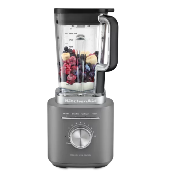Egy szürke KitchenAid turmixgép, tele gyümölcsökkel, mint bogyók és banánszeletek. Az alapja egy sebességszabályzó tárcsával rendelkezik.
