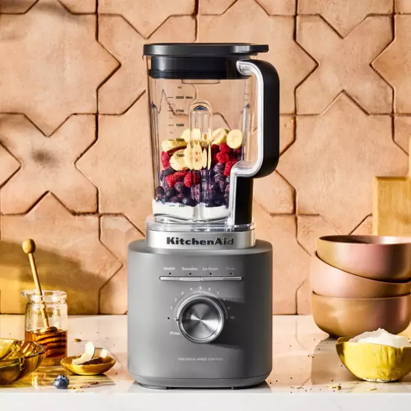 Egy szürke KitchenAid turmixgép bogyókkal és banánszeletekkel egy fehér felületen. Méz és tálak az előtérben.