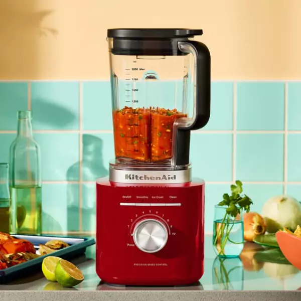 Piros KitchenAid turmixgép salsával. Limák, zöldségek és üvegedények a pulton. Kék és sárga háttér.