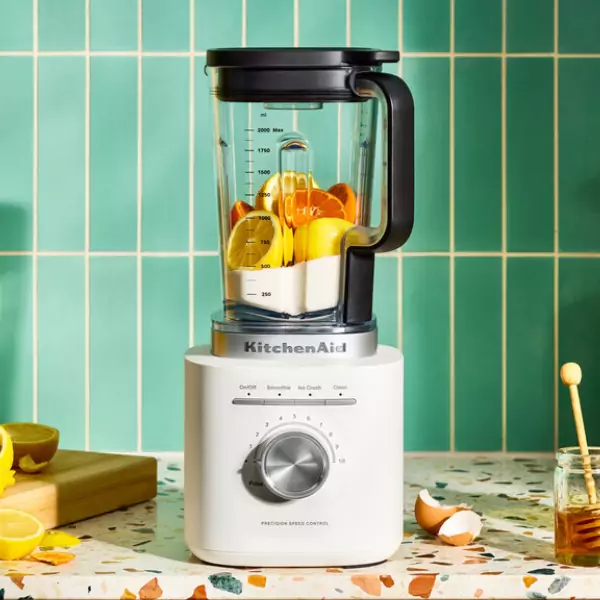 Egy fehér KitchenAid turmixgép gyümölccsel. Egy fa mézcsorgató, egy üveg méz és egy tojáshéj van a közelben.