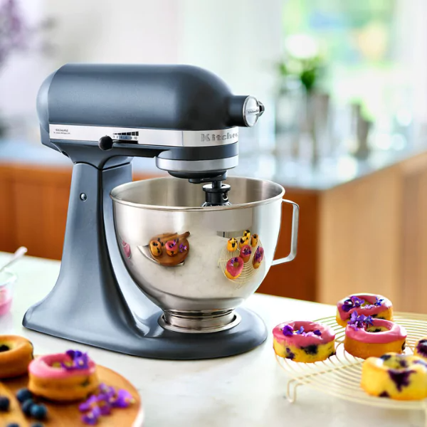 Egy sötétszürke KitchenAid mixer rózsaszín mázas fánkok és virágok mellett. A háttérben egy konyhai környezet látható.