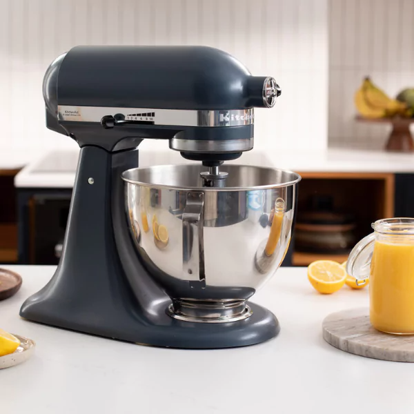 Egy sötétszürke KitchenAid állványos mixer egy fehér pulton áll. Mellette egy pohár lé, citromok és banánok láthatók.