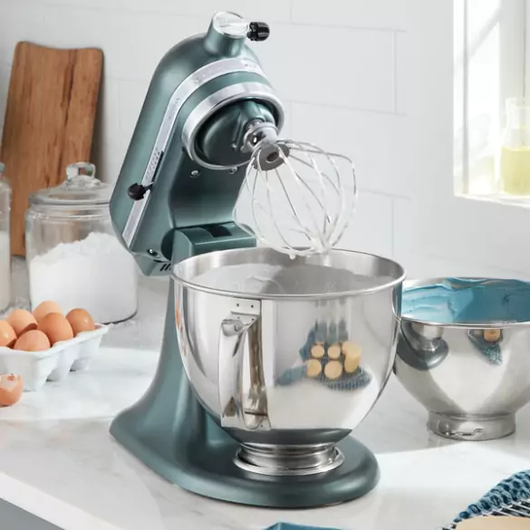 Egy türkiz színű KitchenAid mixer habverő tartozékkal, rozsdamentes acél tál. Tojások, liszt és egy másik tál a háttérben.