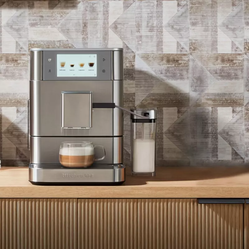 Egy KitchenAid kávéfőző egy pohár kávéval, tejeskannával és fa felülettel. A háttérben geometriai mintázatú fal látható.