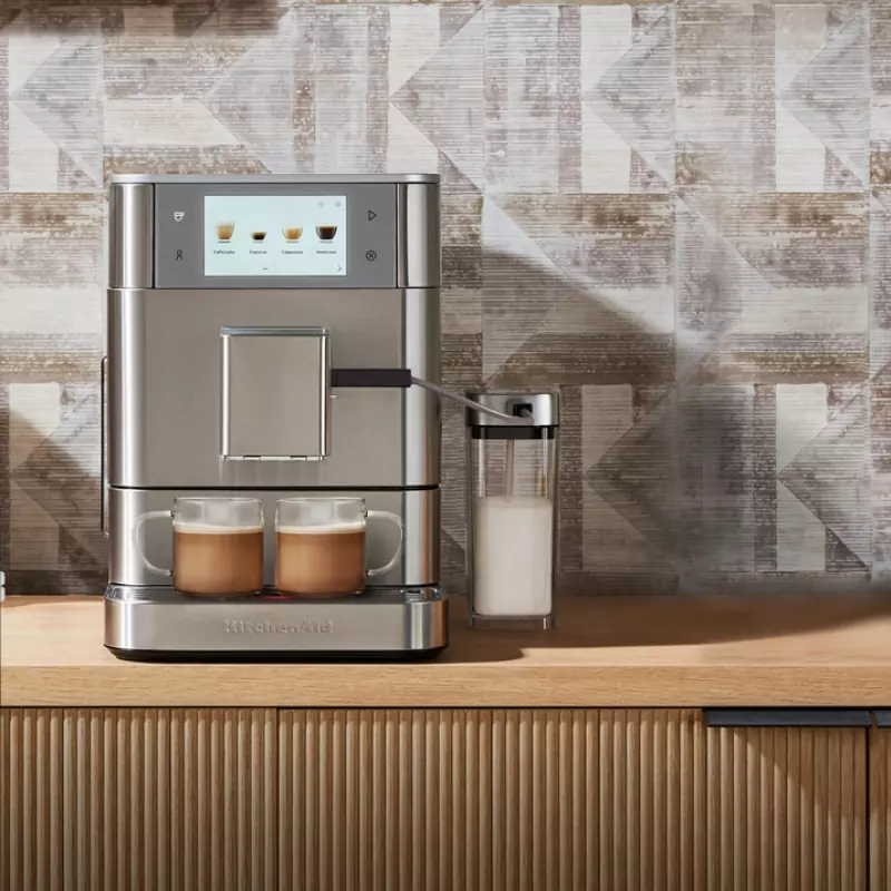 Egy KitchenAid kávéfőző két kávéscsészével, tejhabosítóval és mintás csempével egy fa pulton.