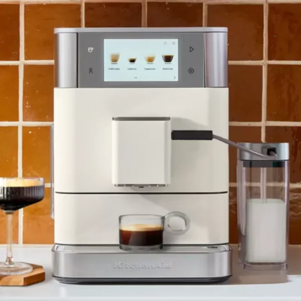 Egy fehér KitchenAid eszpresszógép érintőképernyővel, két kávéscsészével és egy tejhabosítóval. A háttér csempézett.