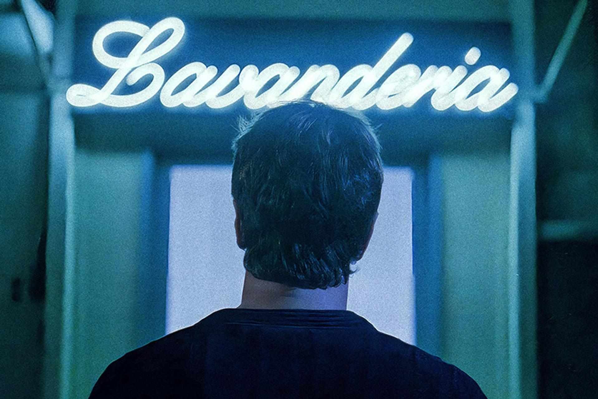 Eine Person mit dem Rücken zum Leuchtschild 'Lavanderia'. Die Szene ist innen und mit kühlem blauem Licht beleuchtet.