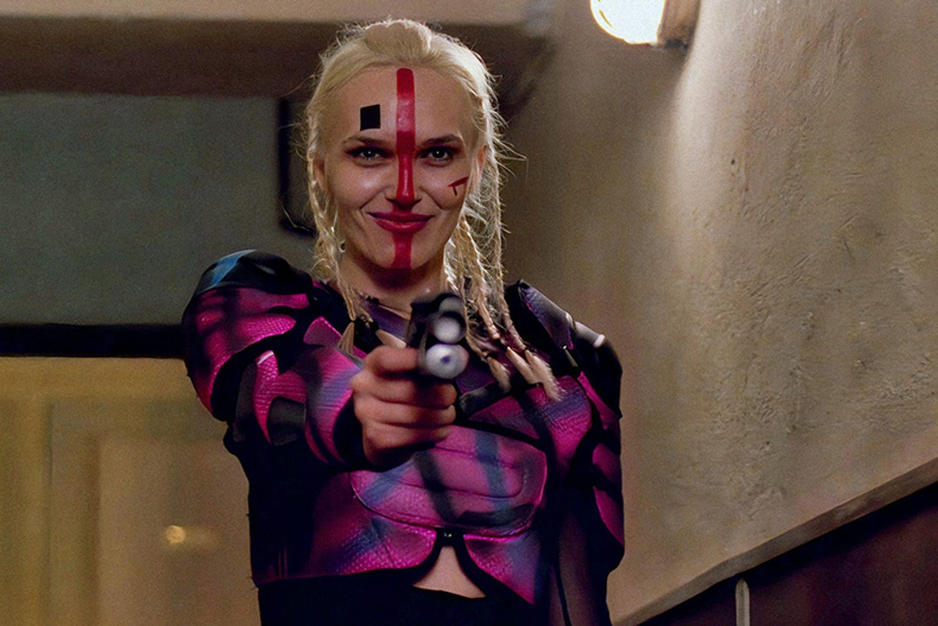 Eine Frau mit Gesichtsbemalung zielt mit einer Waffe. Sie hat blondes Haar, eine pink-schwarze Jacke und lächelt. Der Hintergrund ist ein Flur.