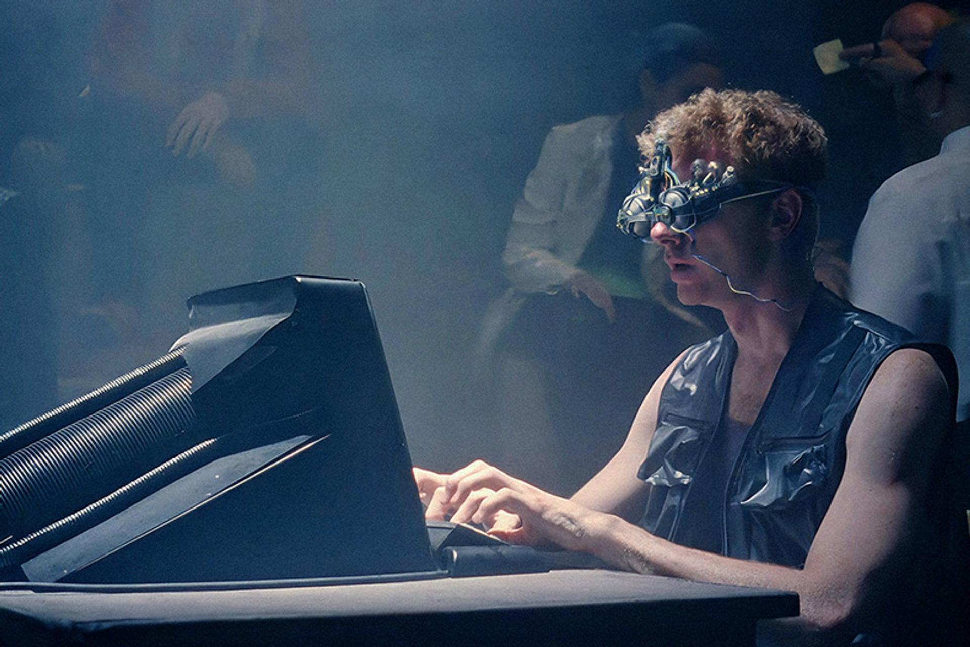 Ein Mann mit einem verdrahteten Gerät auf dem Kopf tippt an einem Computer. Andere Figuren sind im Hintergrund, in einem dunklen, rauchigen Raum.