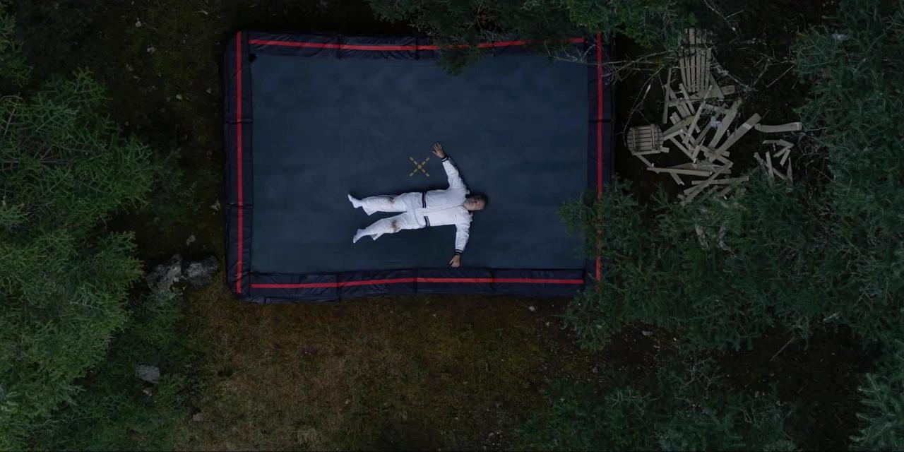 Draufsicht: Eine Person in weißem Anzug liegt auf einem Trampolin, umgeben von Wald. Trümmer befinden sich im Hintergrund.