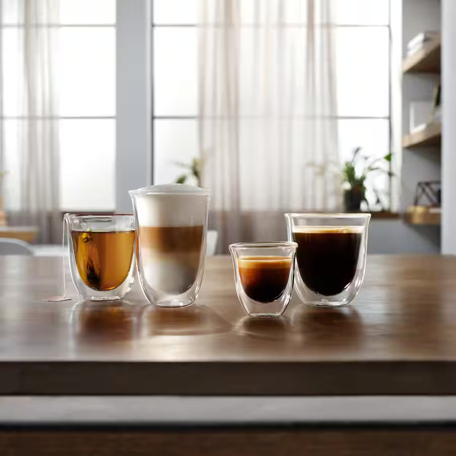 Cztery szklanki z napojami: herbatą, latte, espresso i kawą. Stoją na drewnianym stole przed oknem.