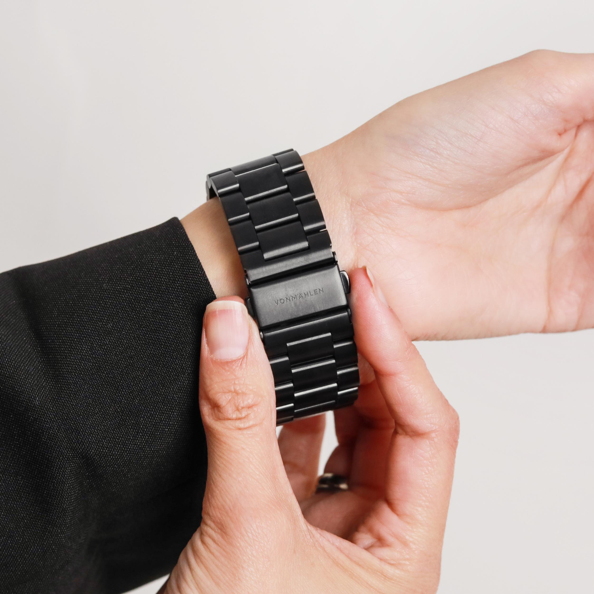 Handgelenk einer Person mit einer schwarzen Metalluhr. Die Uhr wird mit der anderen Hand geschlossen.