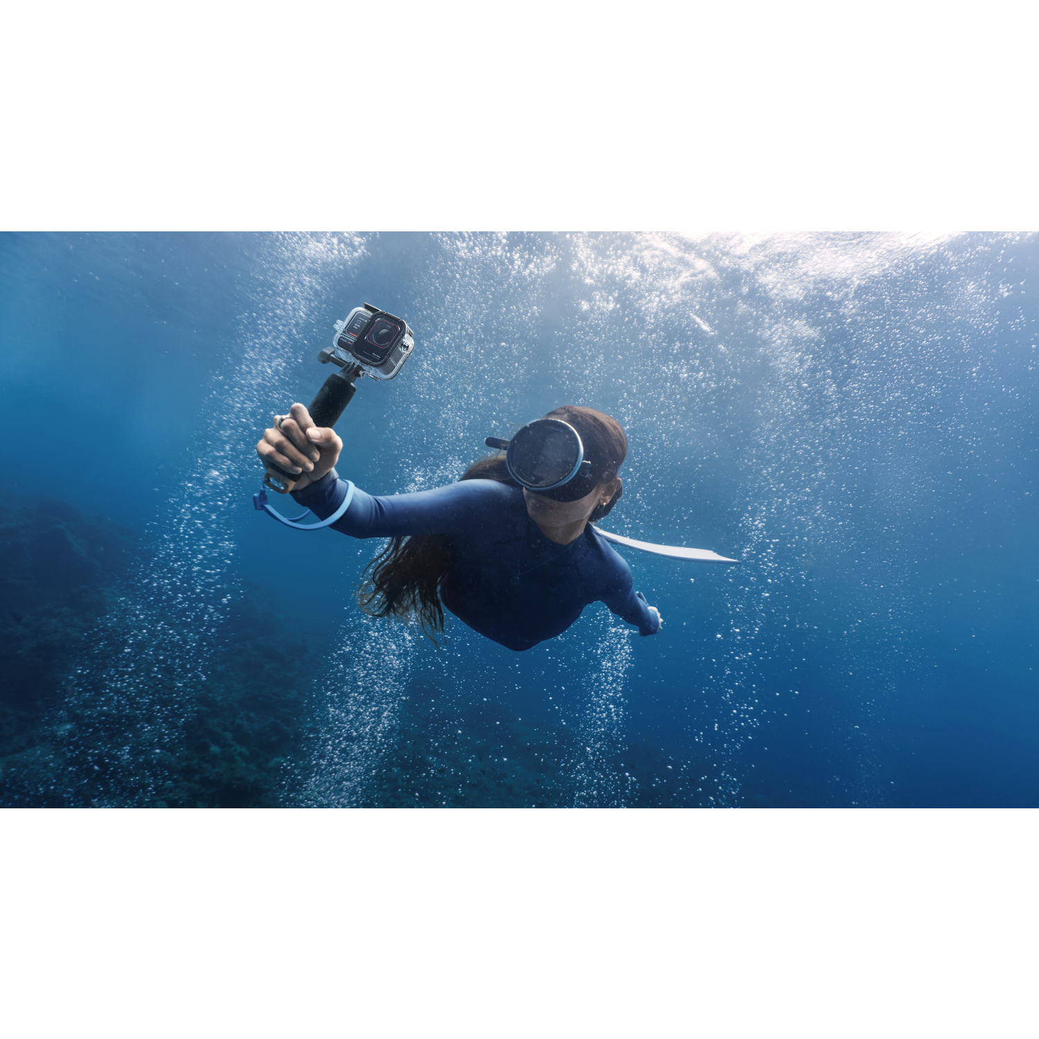 A person dives underwater, holding a camera. Bubbles rise. The diver wears a wetsuit, mask, and snorkel, with a camera in hand.