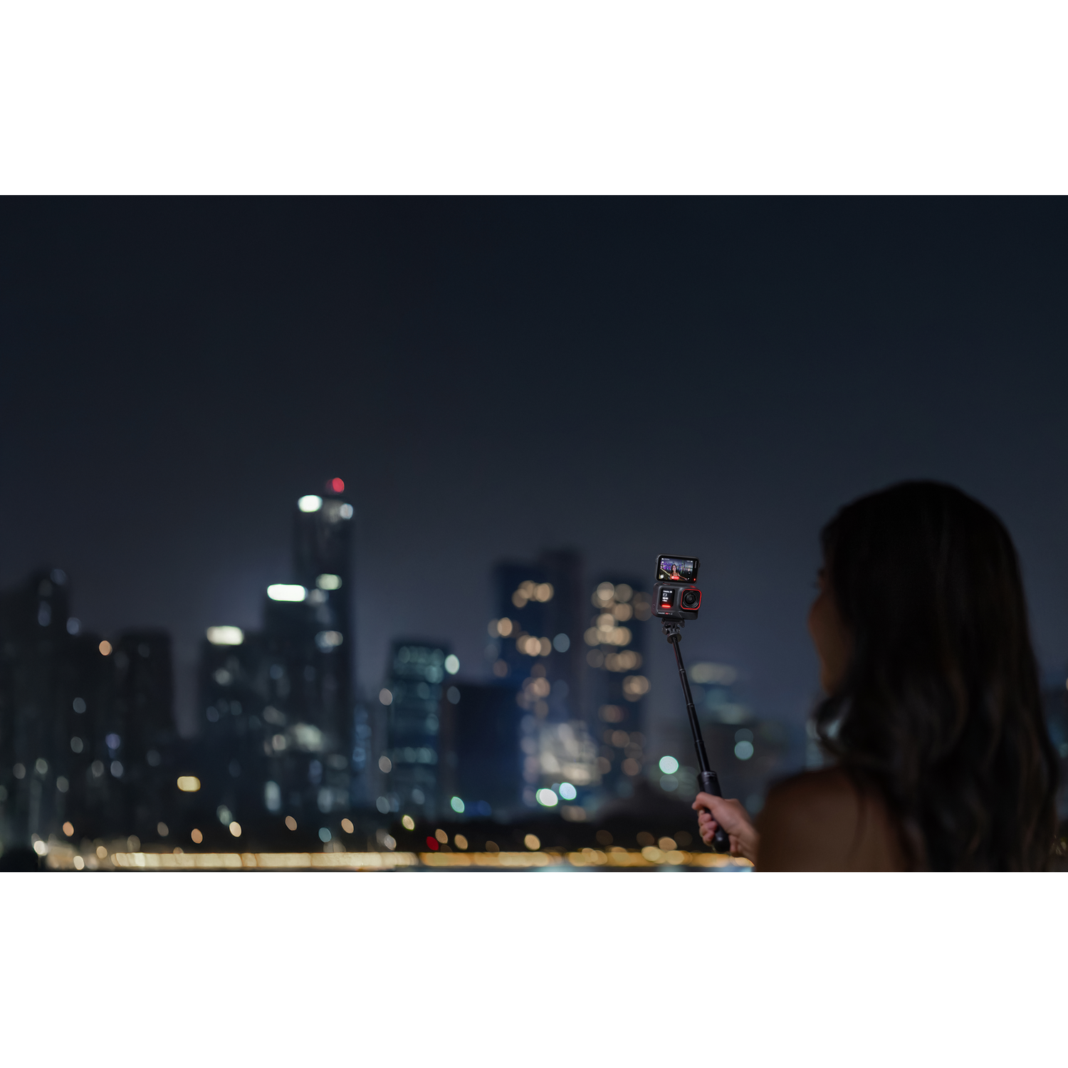 A person holds a selfie stick with a camera, taking a photo at night with city lights in the background.