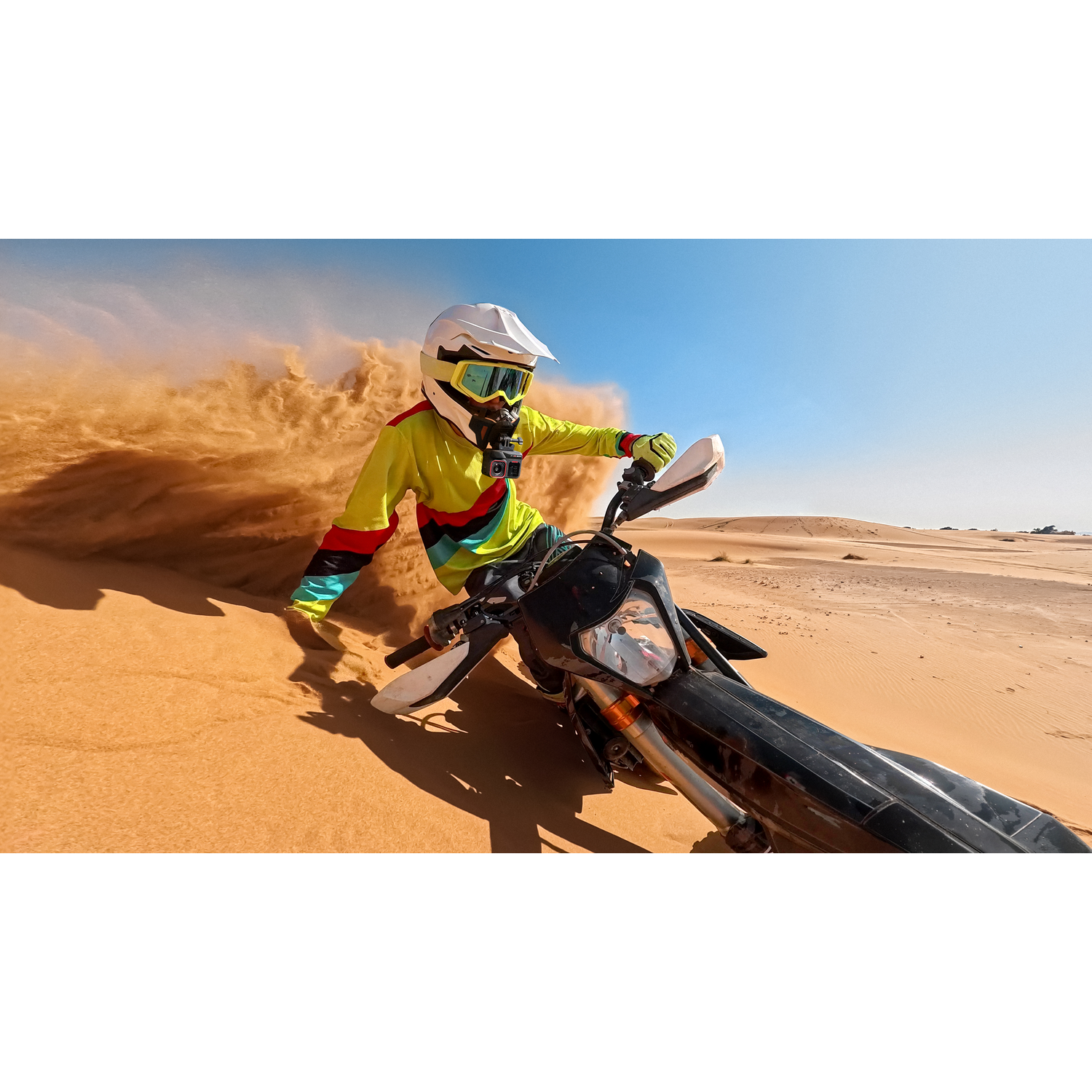 A person on a motorcycle in a desert, kicking up sand. Blue sky in the background. The person wears a helmet and colorful clothing.