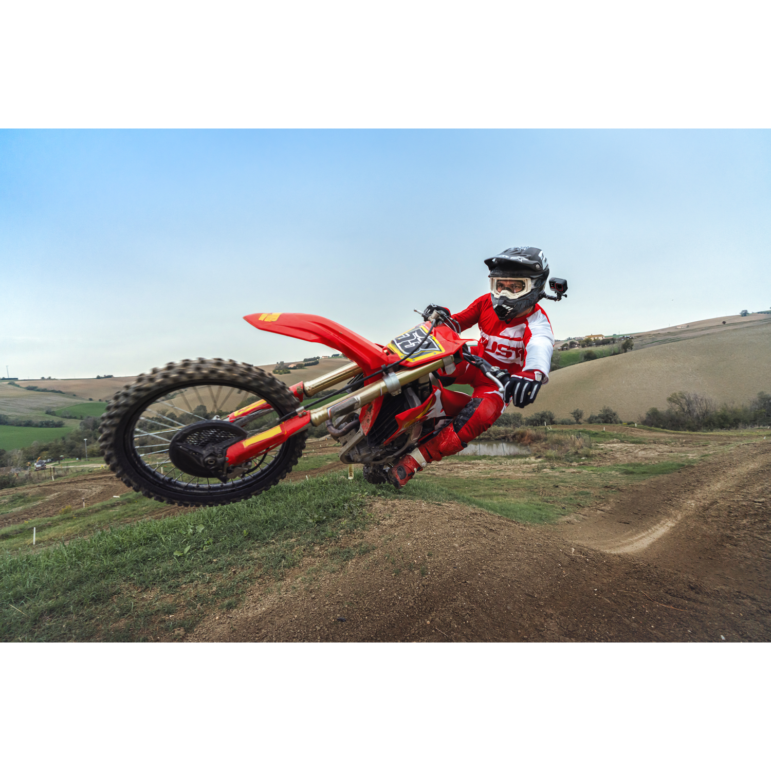 A motocross rider in red gear jumps a dirt mound. The red bike is mid-air. Green fields and a blue sky form the background.