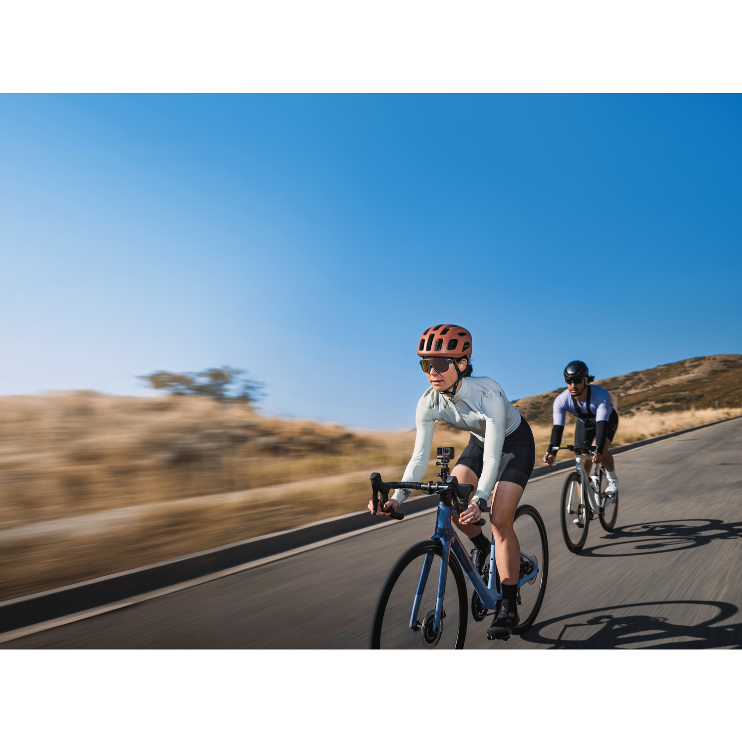 Two cyclists ride uphill on a paved road. The lead cyclist wears a helmet and light clothing. Clear blue sky in the background.