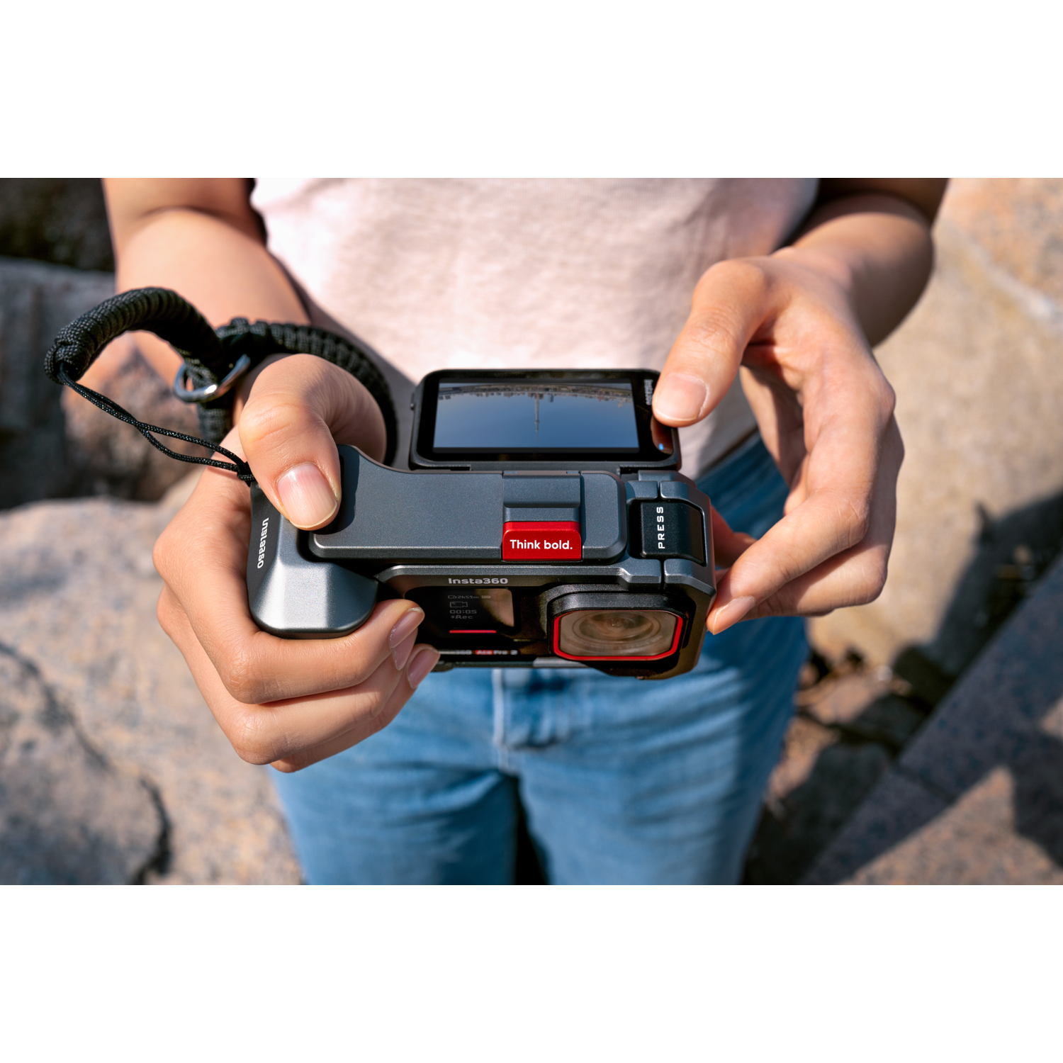 Une personne tient une caméra Insta360 grise, montrant un écran et un objectif. La caméra a un bouton rouge avec l'inscription 'Think bold'.