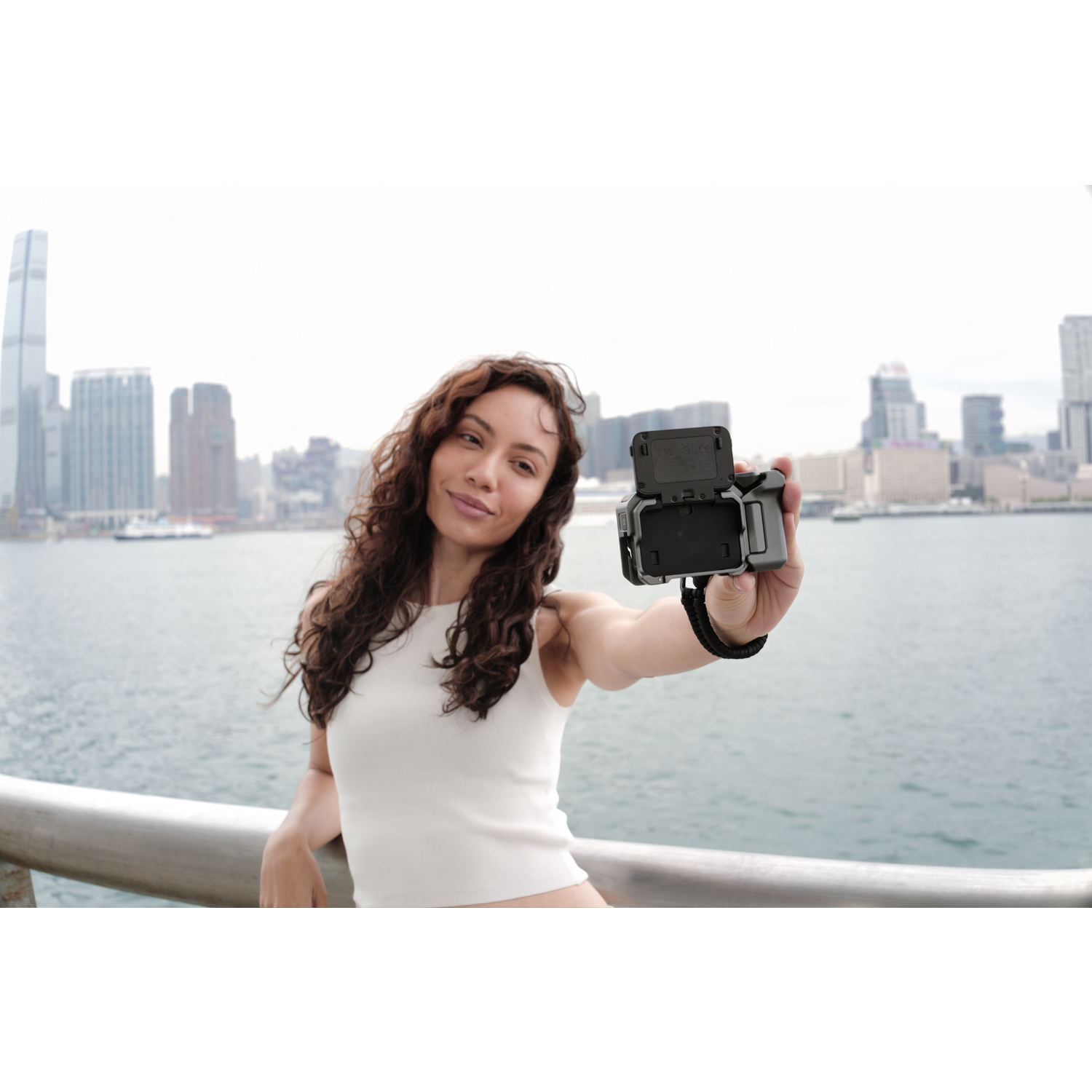 Une femme prend un selfie avec une caméra, souriante. Paysage urbain et eau en arrière-plan.