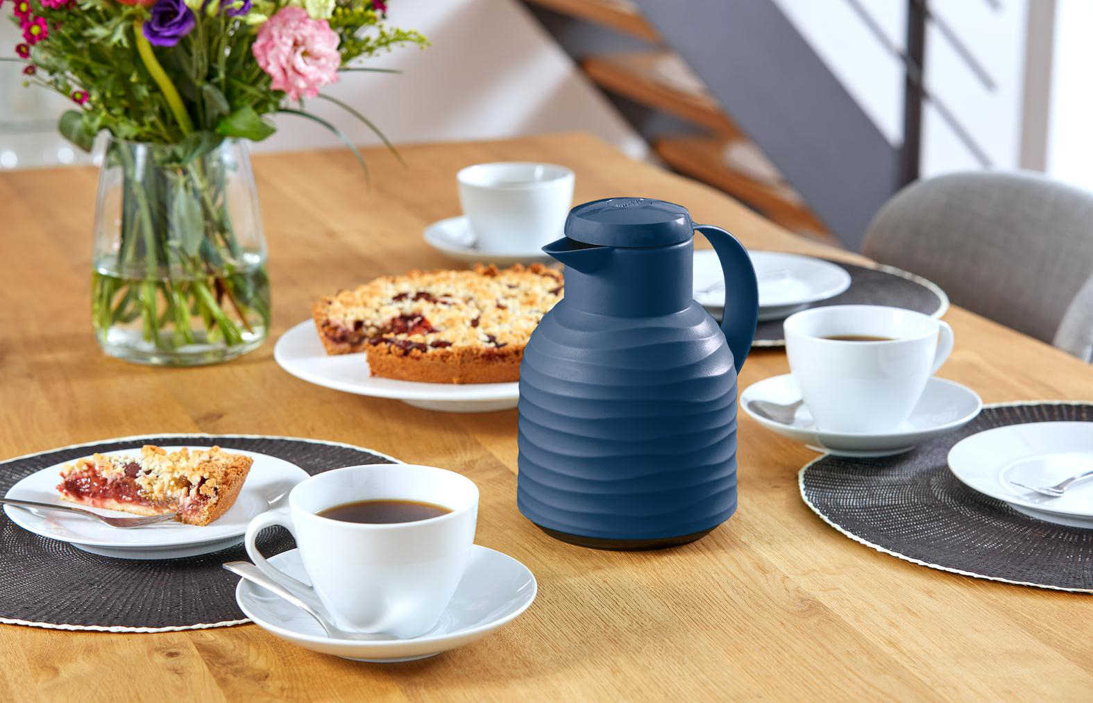 Ein Tisch ist mit Kaffee, Kuchen und einer blauen Thermoskanne gedeckt. Links steht eine Blumenvase.