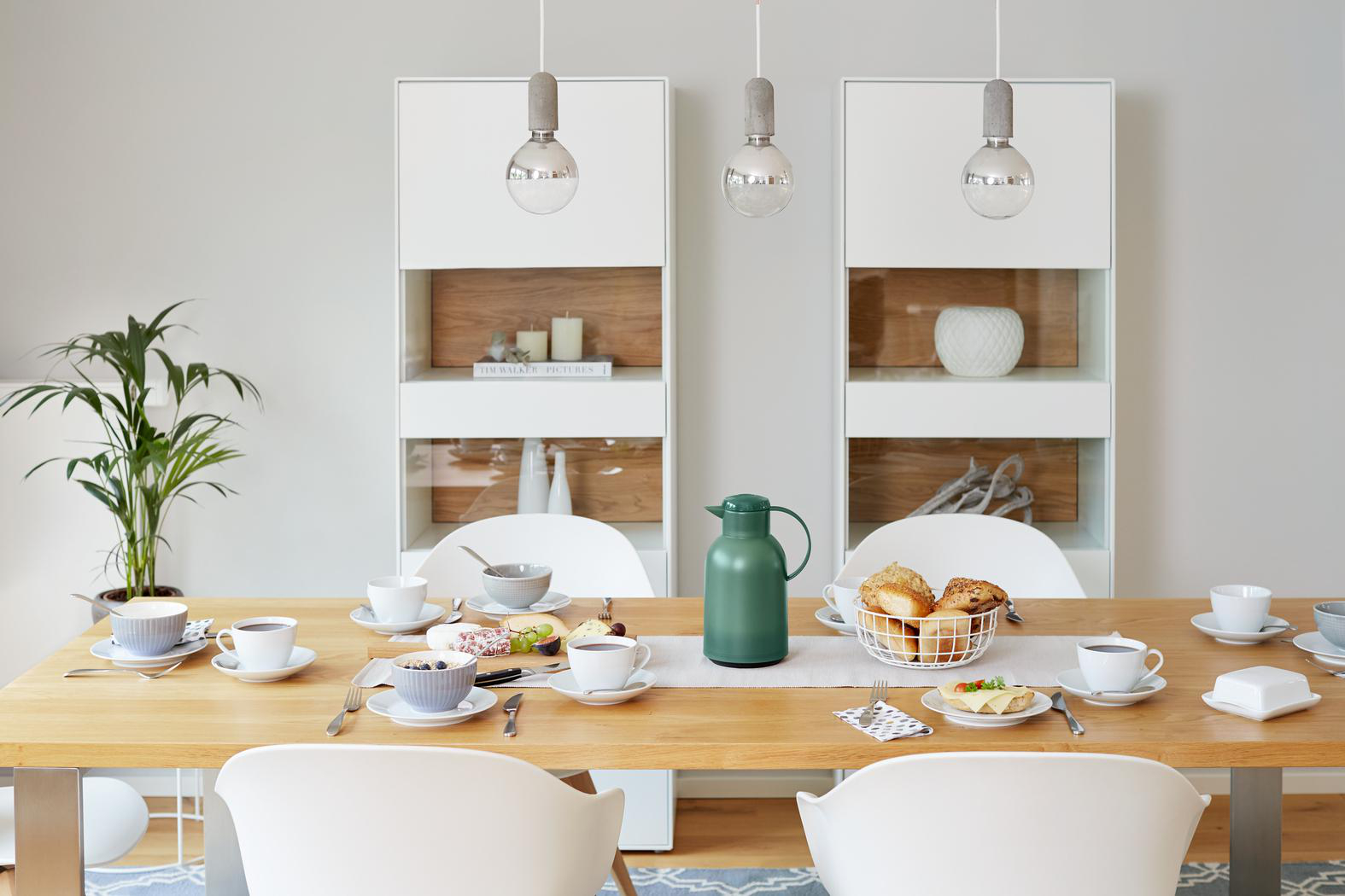 Ein gedeckter Tisch für das Frühstück mit Kaffee, Brot und Obst. Schränke und Hängelampen sind im Hintergrund.