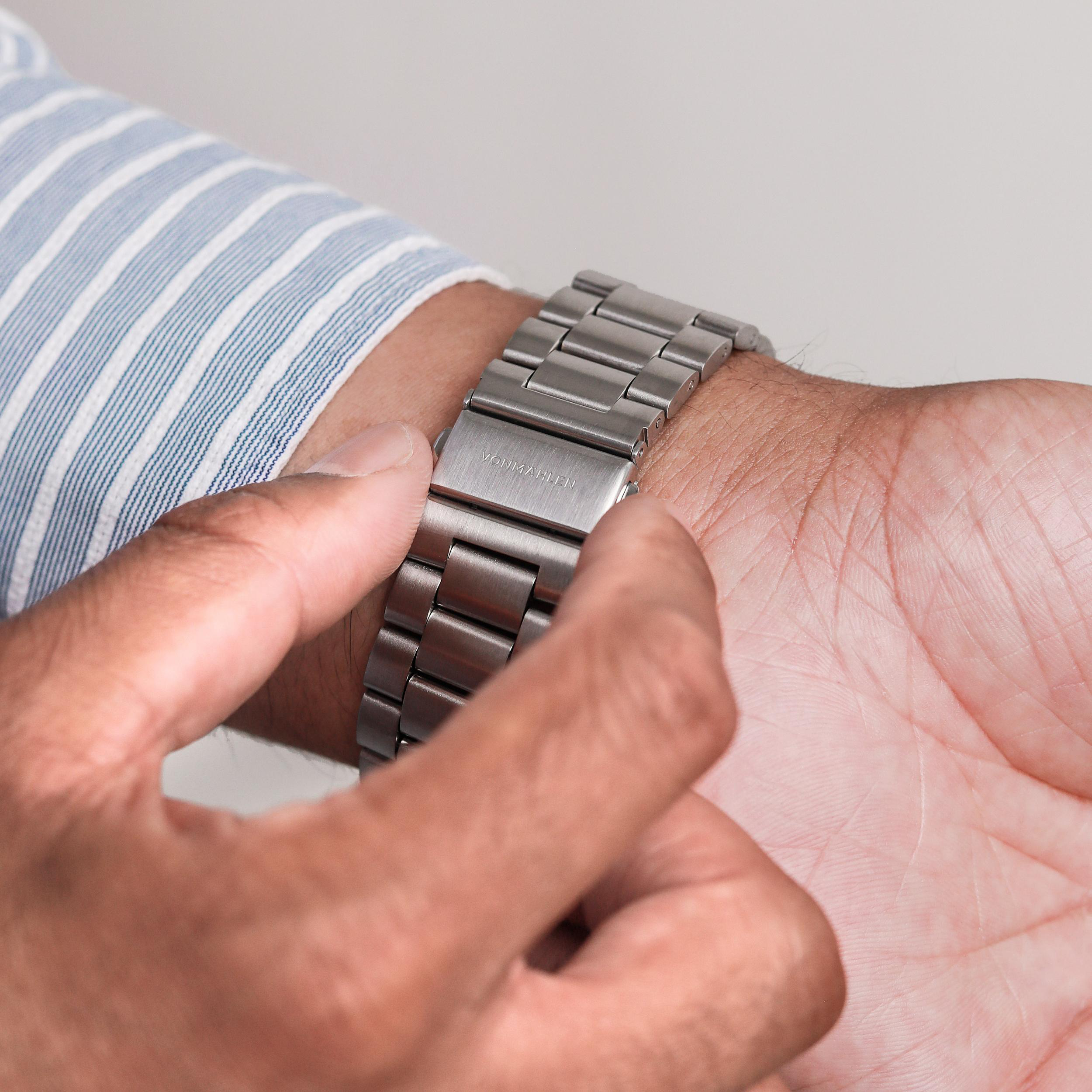 Eine Person stellt eine silberne Uhr an ihrem Handgelenk ein. Das Uhrenarmband ist aus Metall. Das Hemd ist blau und weiß gestreift.