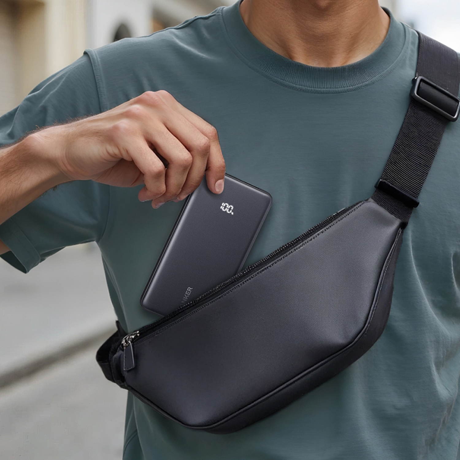 A person in a teal shirt is holding a power bank, taking it out of a black sling bag. The power bank shows 100%.