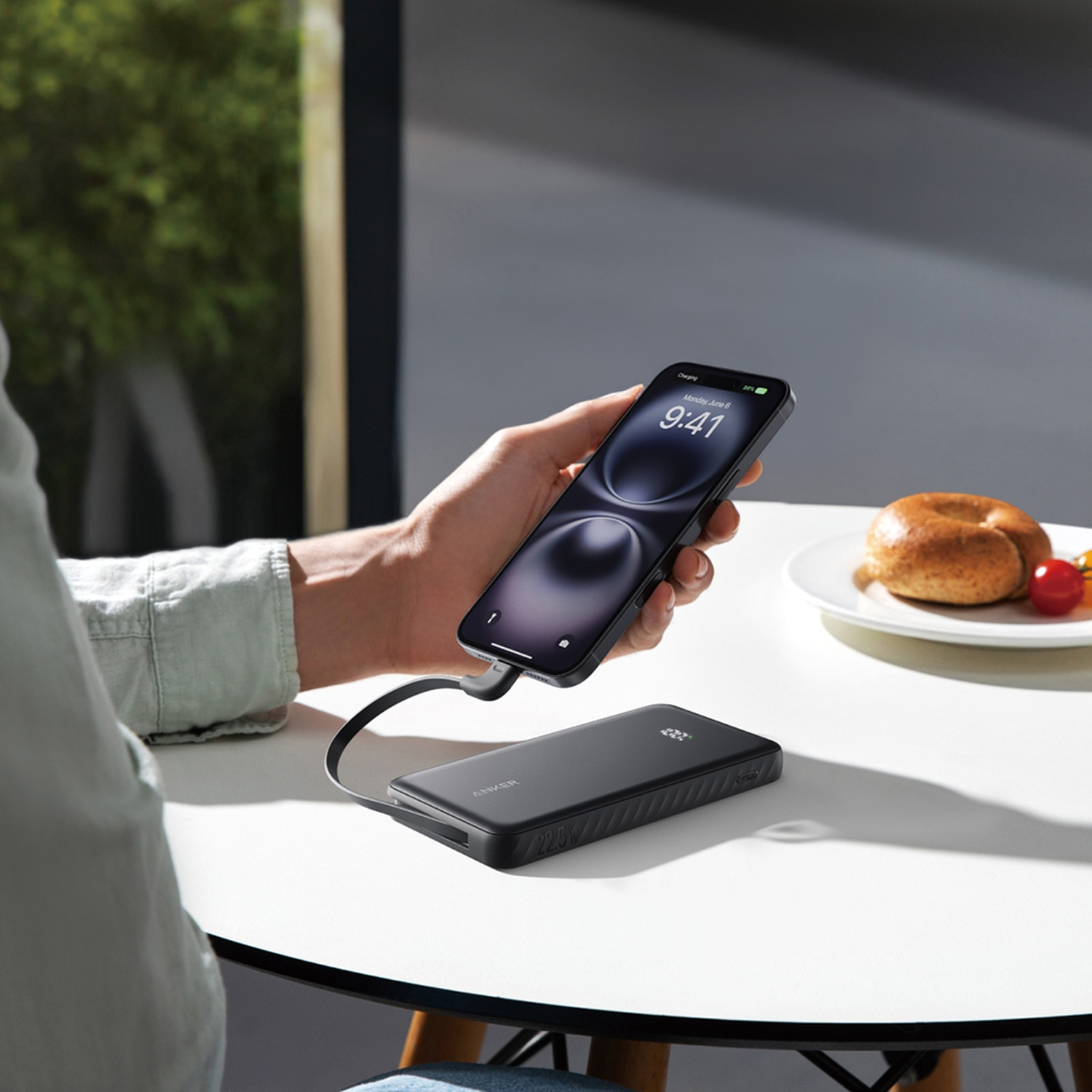 Une main tient un téléphone en chargeant à partir d'une batterie externe Anker noire sur une table blanche. Un bagel et des tomates sont sur une assiette.