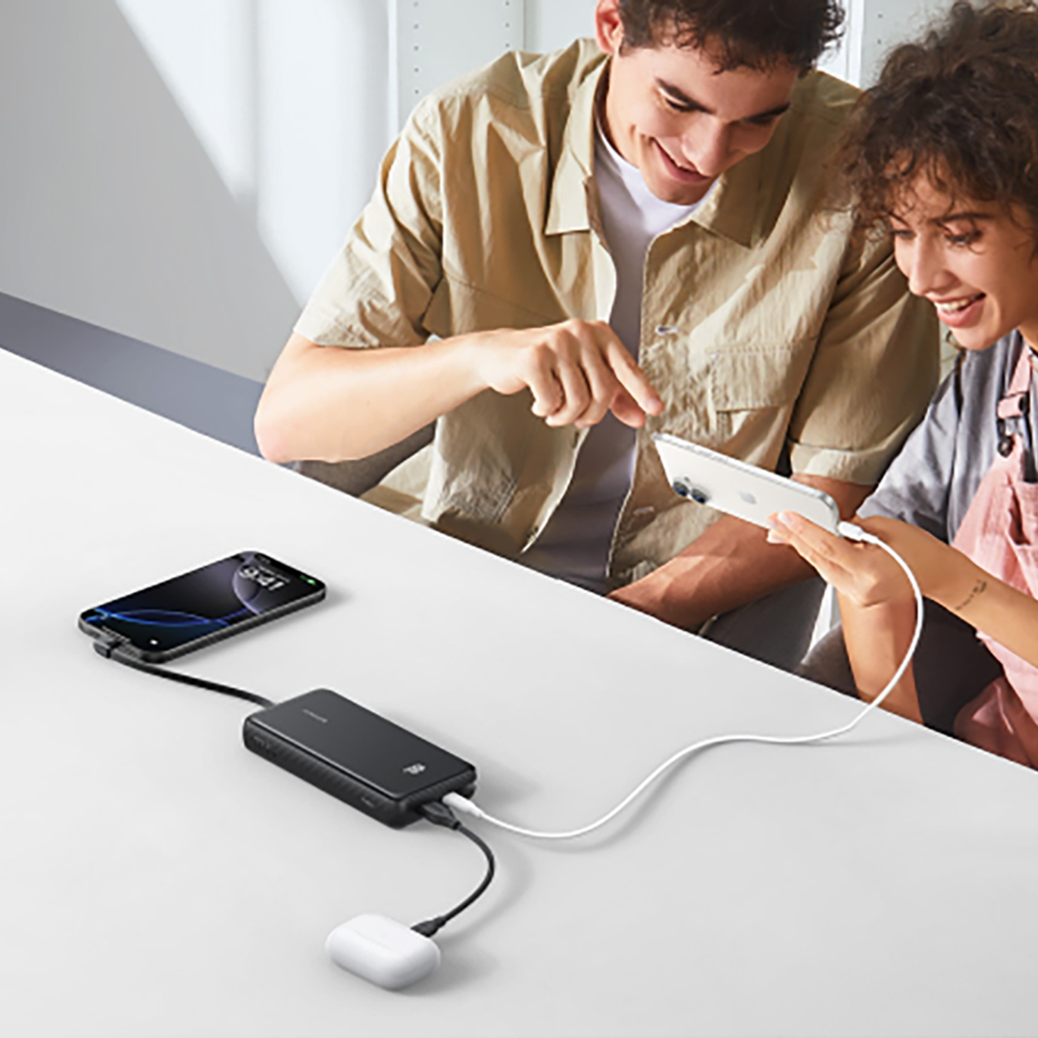 Deux personnes regardent des téléphones connectés à une batterie externe. Un téléphone et des écouteurs sont également connectés. Table blanche.