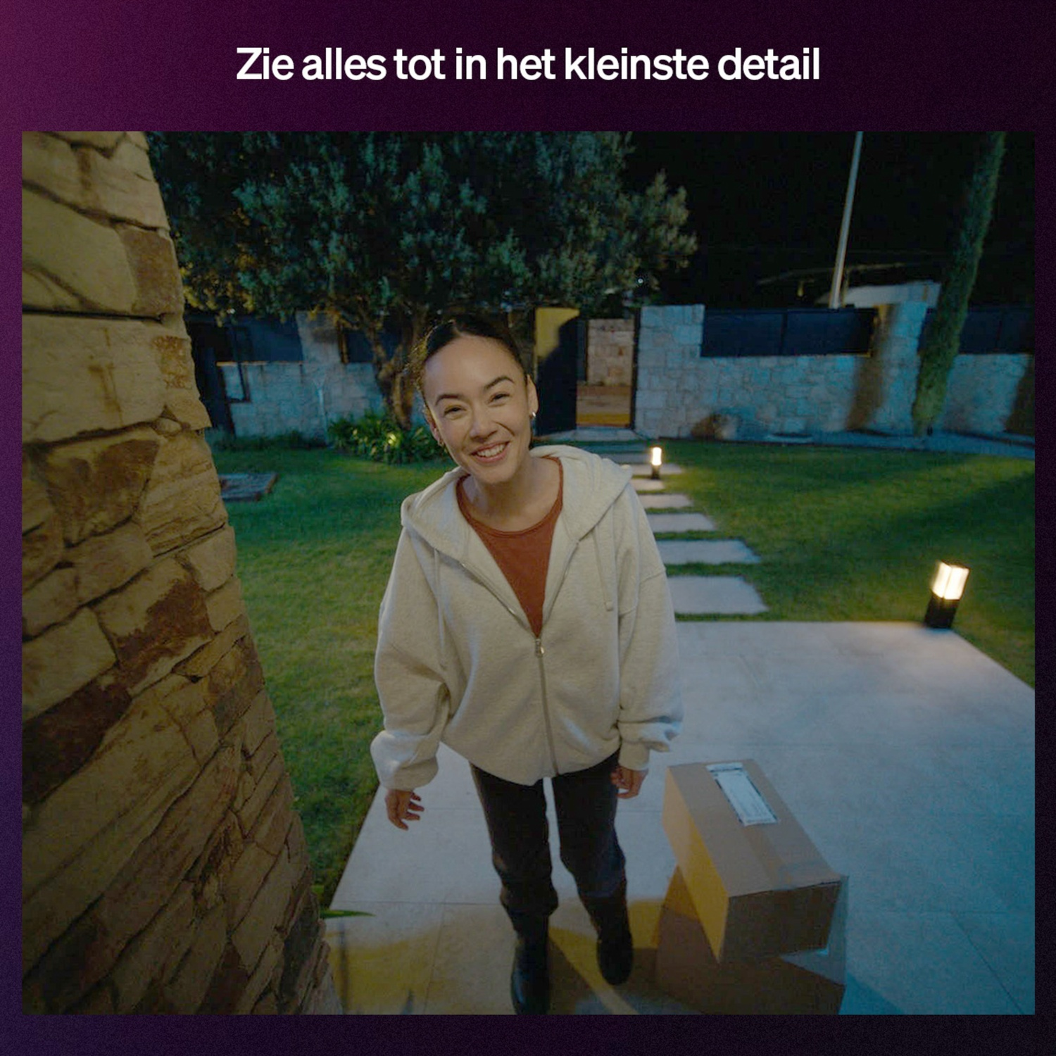 Woman smiles, standing near a box on a porch at night. Stone wall and green grass in the background. Lit pathway.