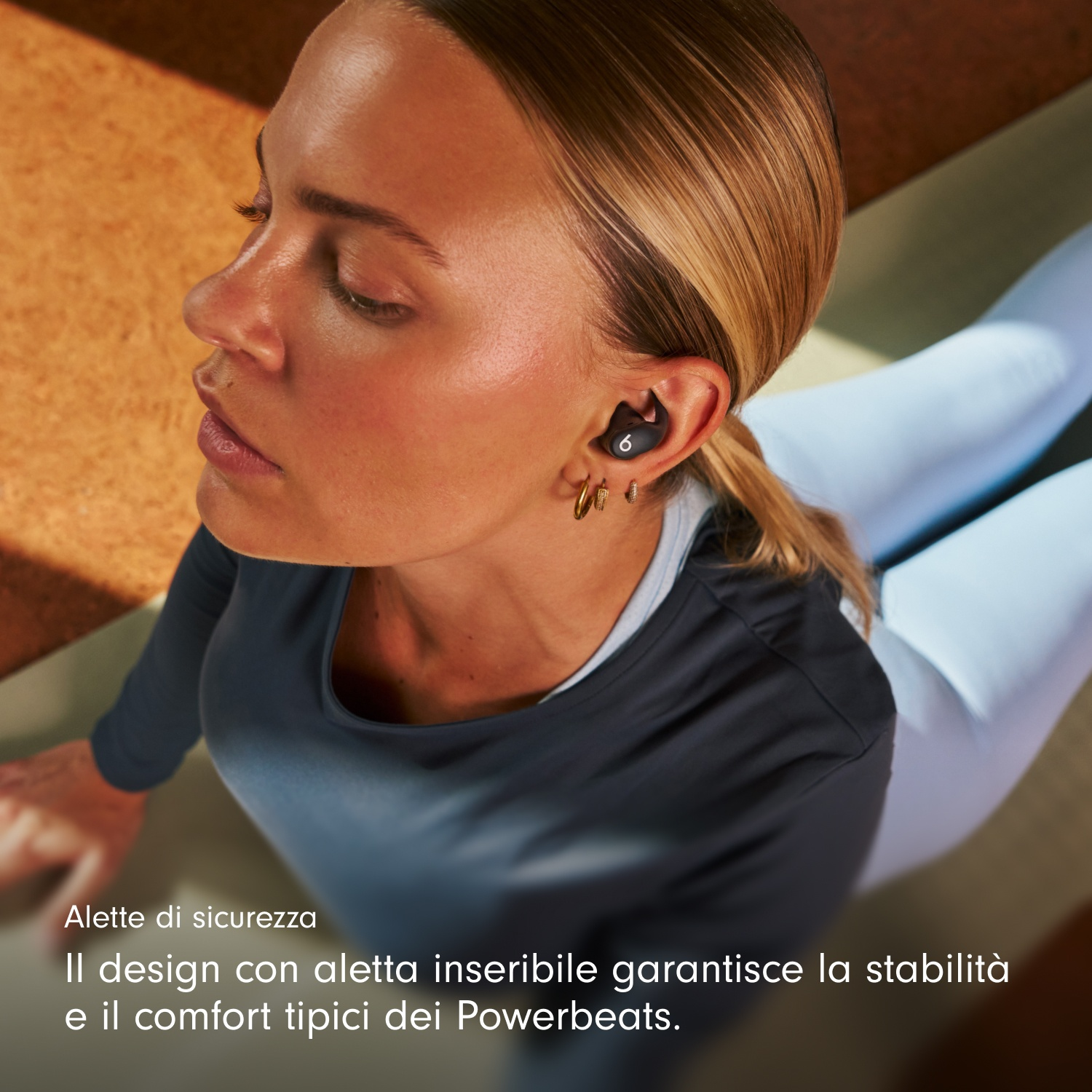 Una donna con auricolari è sdraiata. Ha i capelli biondi e indossa una maglia scura e leggings azzurri. Lo sfondo è sfocato.