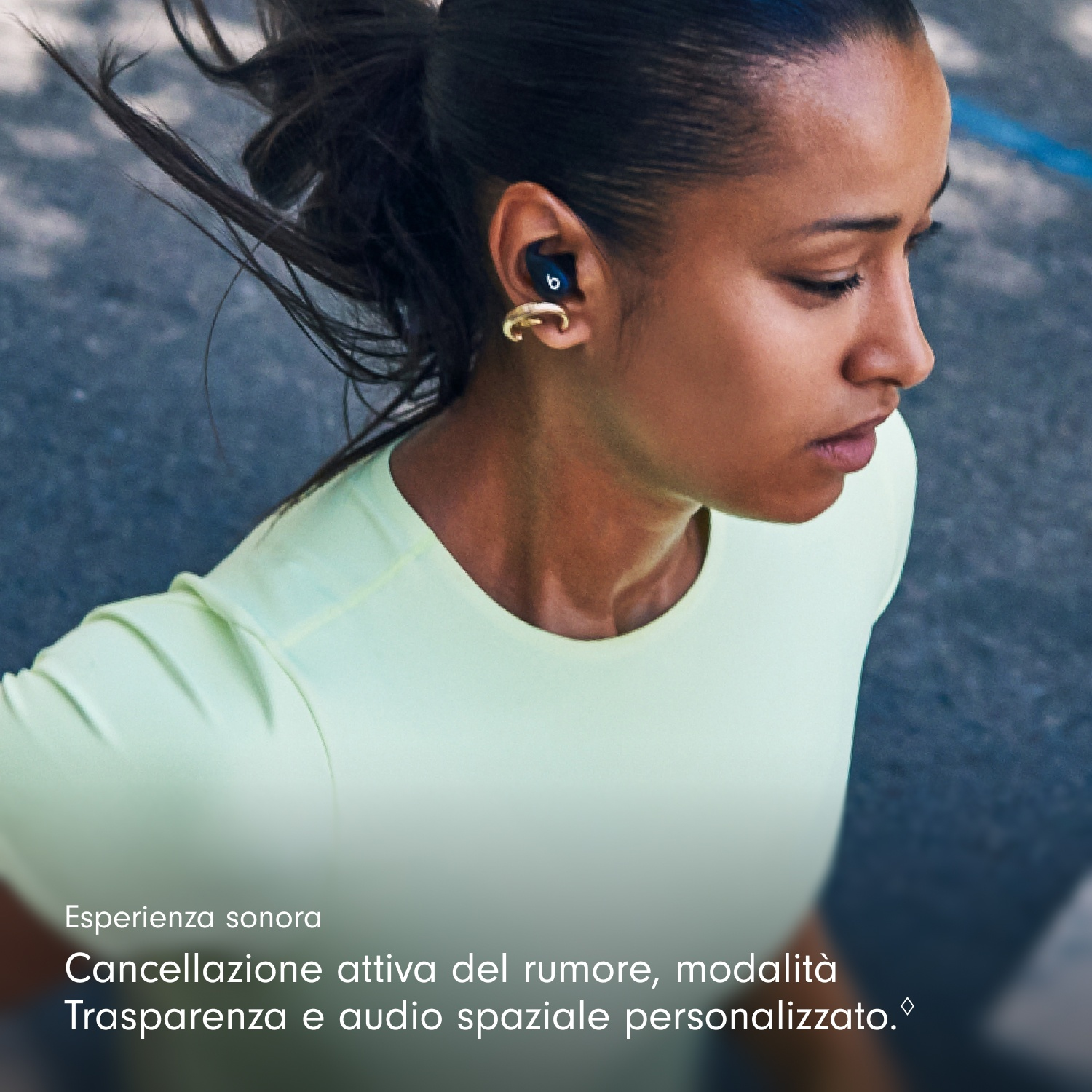 Una donna con i capelli scuri che indossa auricolari, orecchino d'oro e camicia verde chiaro, che guarda in basso. Lo sfondo è sfocato.