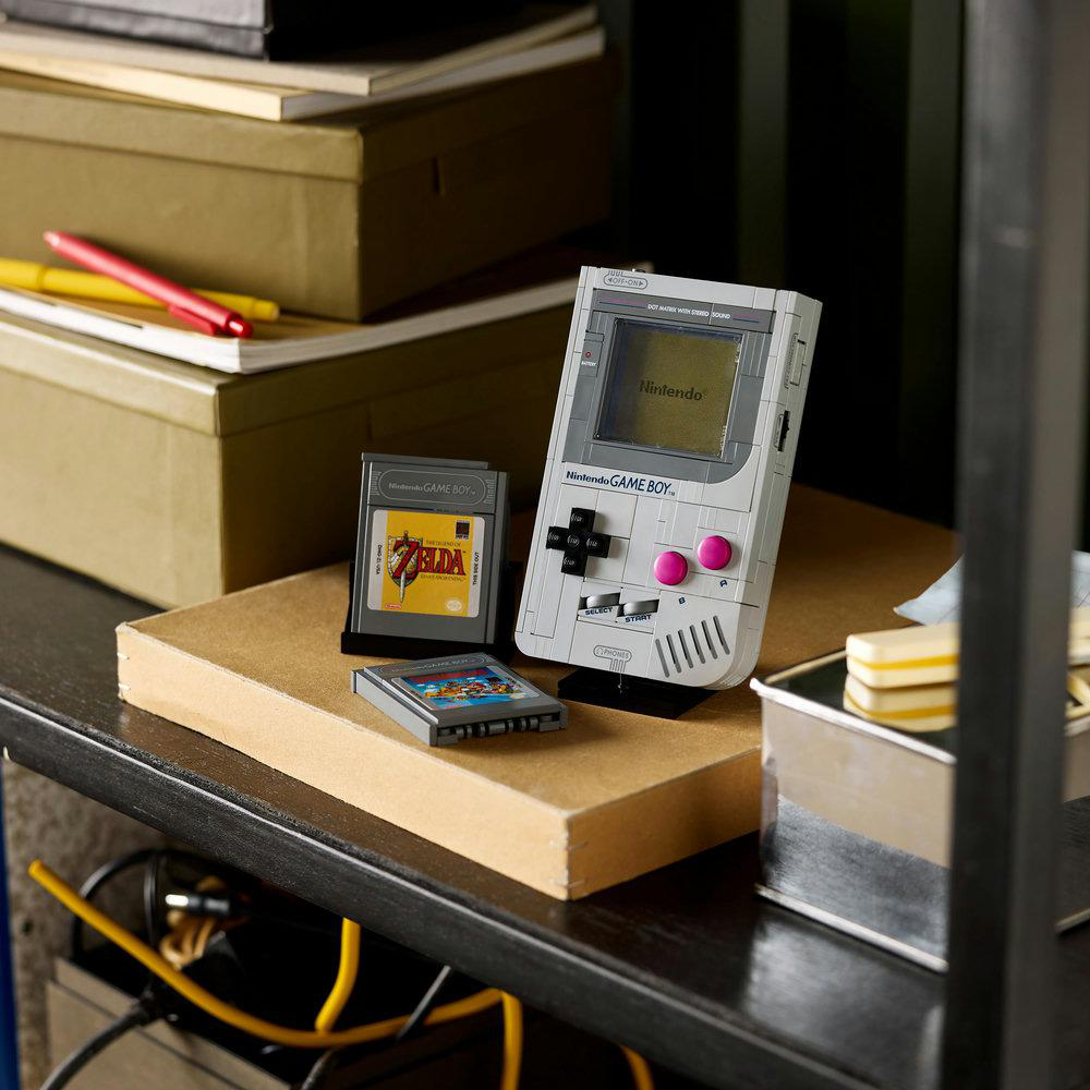 Ein LEGO Game Boy mit Cartridges auf einem Regal. Bücher und Kisten befinden sich im Hintergrund. Stift und gelber Stift liegen auf den Kisten.