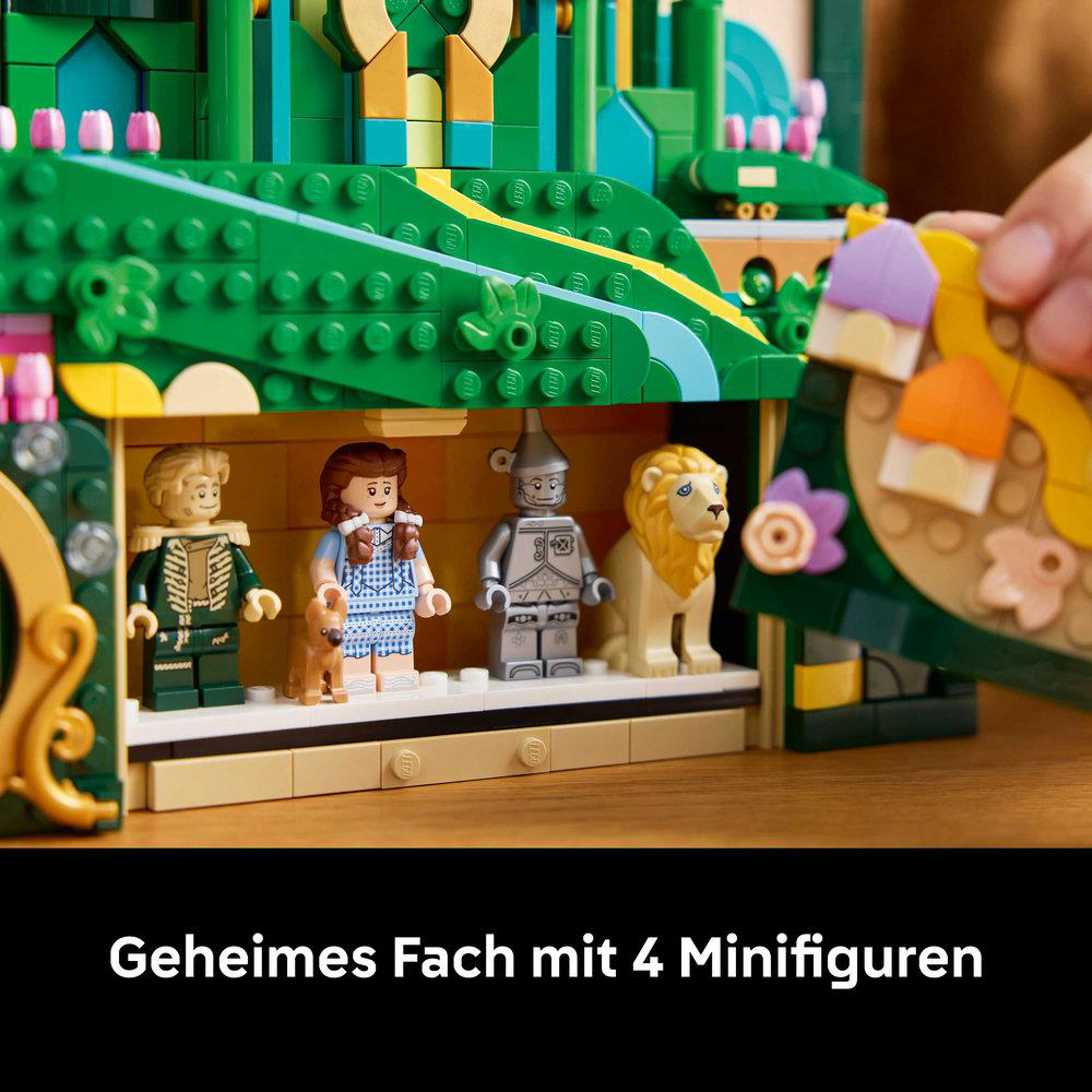 Lego-Set mit 4 Minifiguren: Vogelscheuche, Dorothy, Blechmann und Löwe. Grüne Landschaft im Hintergrund, Hand hält ein Stück.