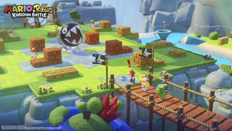 Spielszene: Mario- und Rabbids-Charaktere auf einer Brücke, Chain Chomp, Backsteinbauten, Flaggen und ein Fluss in einer farbenfrohen Umgebung.