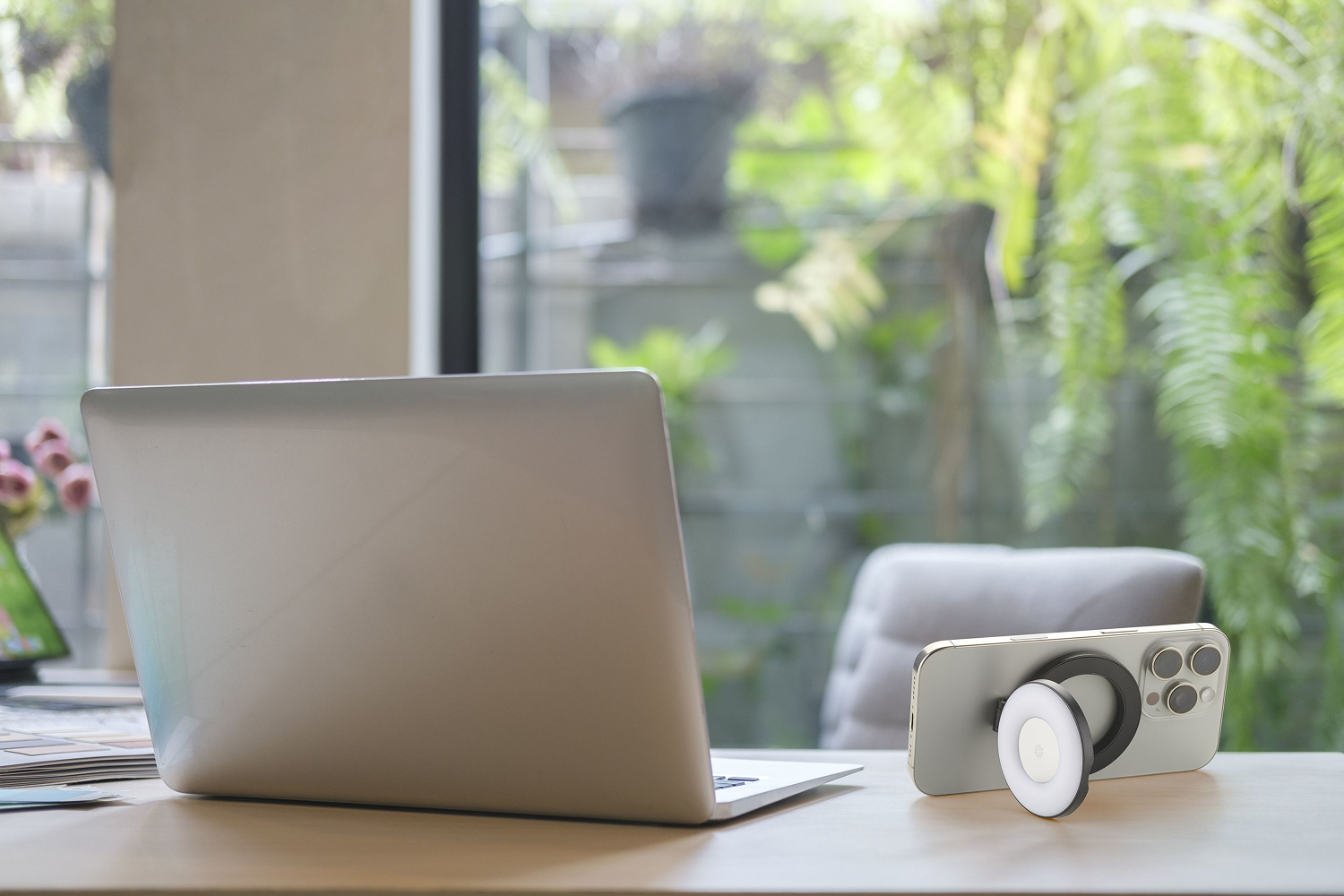 Una laptop y un teléfono con una luz de anillo están sobre un escritorio de madera. Se ven plantas verdes a través de una ventana en el fondo.
