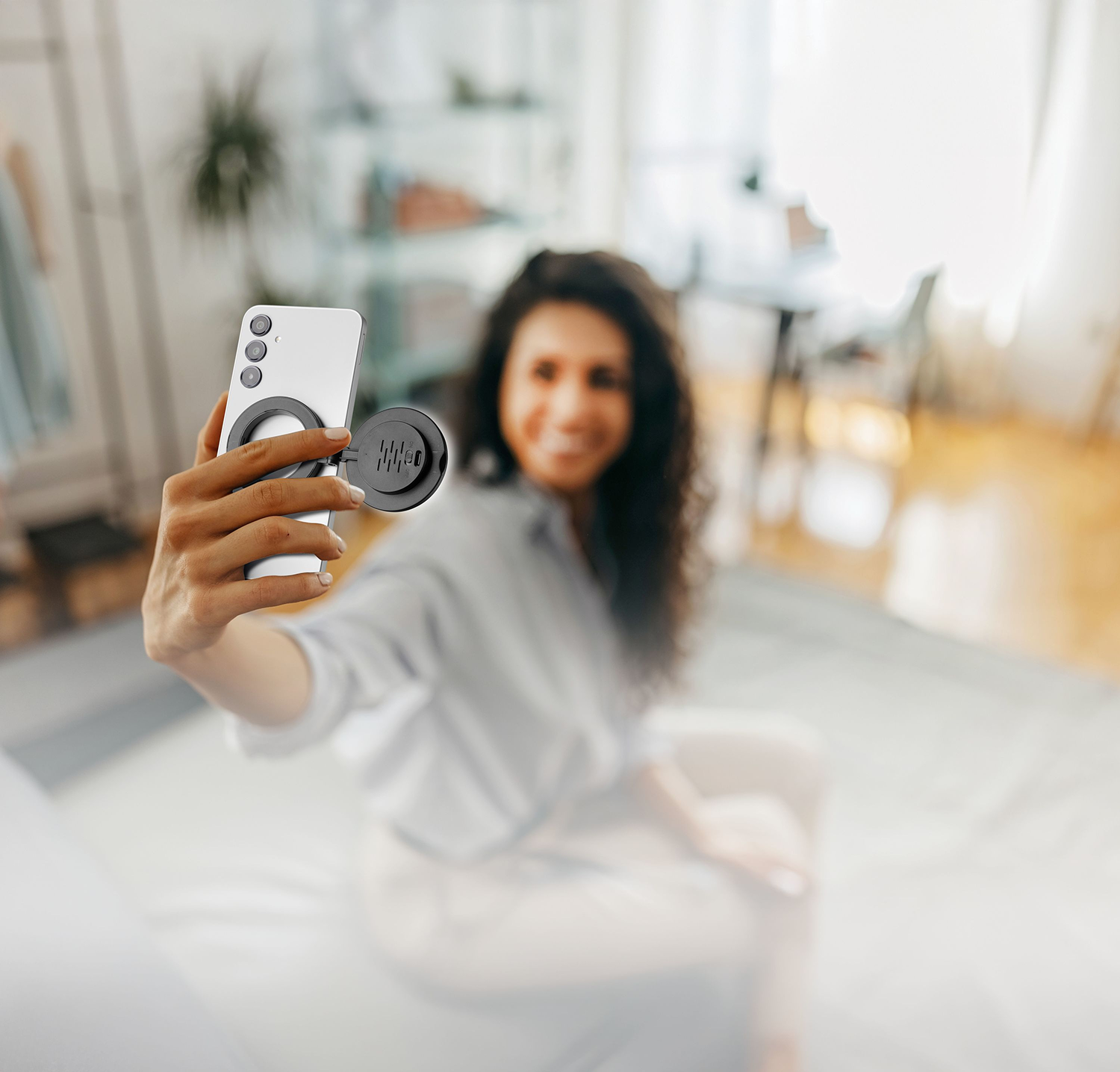 Mujer tomándose una selfie con un teléfono y un accesorio redondo. El fondo incluye una habitación con muebles y luz natural.