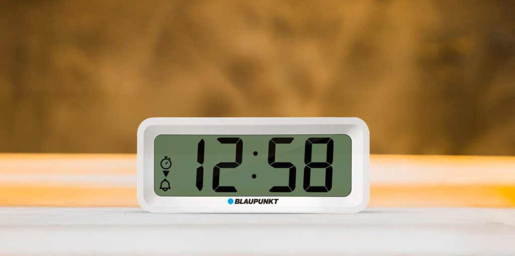 Biały cyfrowy zegar Blaupunkt pokazuje 12:58 na zielonym ekranie. Widoczna ikona timera i dzwonka. Tło jest rozmyte brązowe.