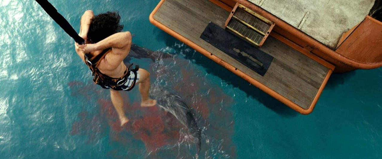 Ein Mann hängt an einem Seil über blutigem Wasser in der Nähe eines Bootes. Ein Hai ist unten sichtbar. Die Szene deutet auf Gefahr hin.