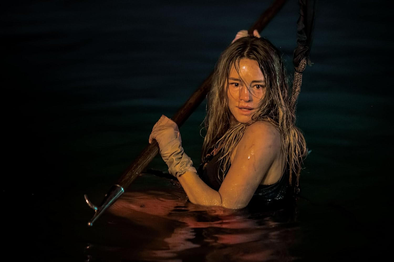 Eine Frau im Wasser hält eine Stange. Sie hat nasse blonde Haare und einen besorgten Gesichtsausdruck. Die Szene ist dunkel.