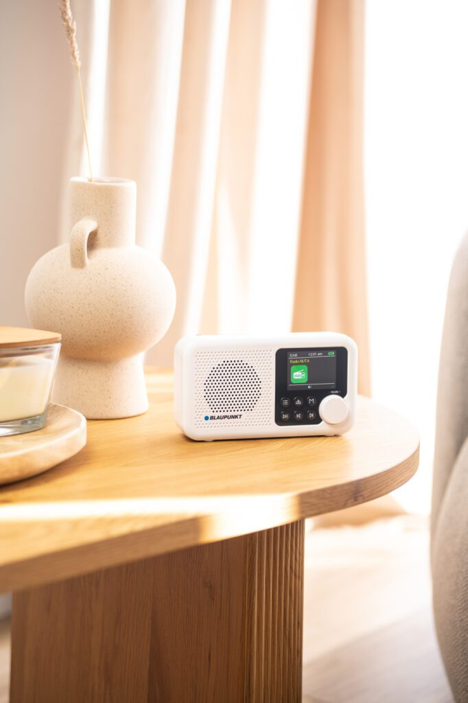 Białe radio stoi na drewnianym stole obok wazonu i świecy. W tle zasłona.