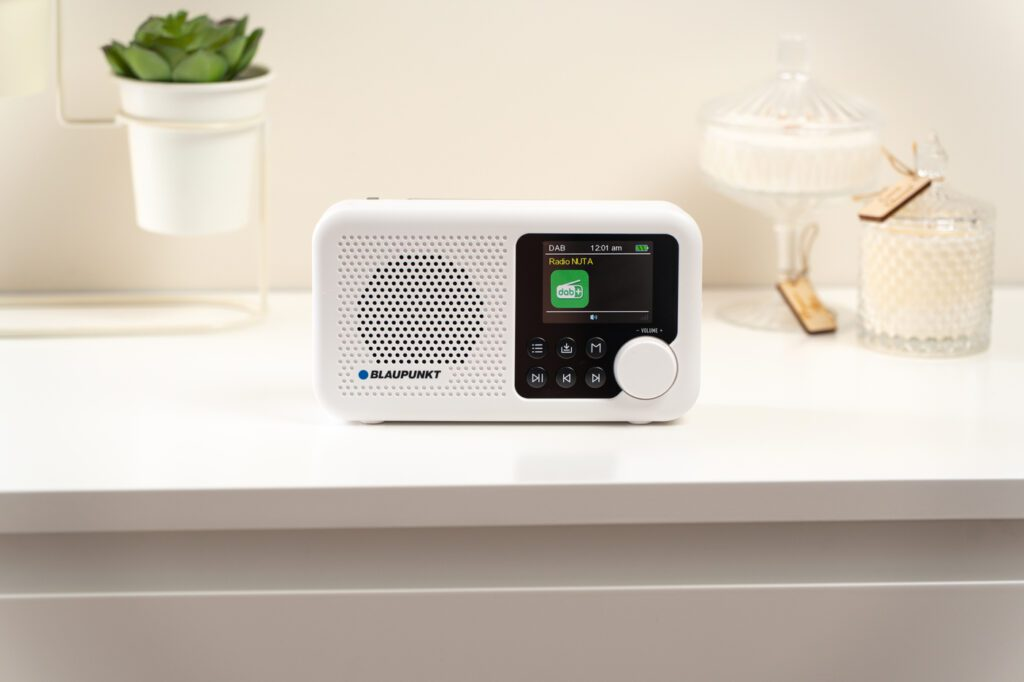 Białe radio Blaupunkt stoi na białej powierzchni. Mała zielona roślina w białej doniczce po lewej. Szklane dekoracje po prawej.