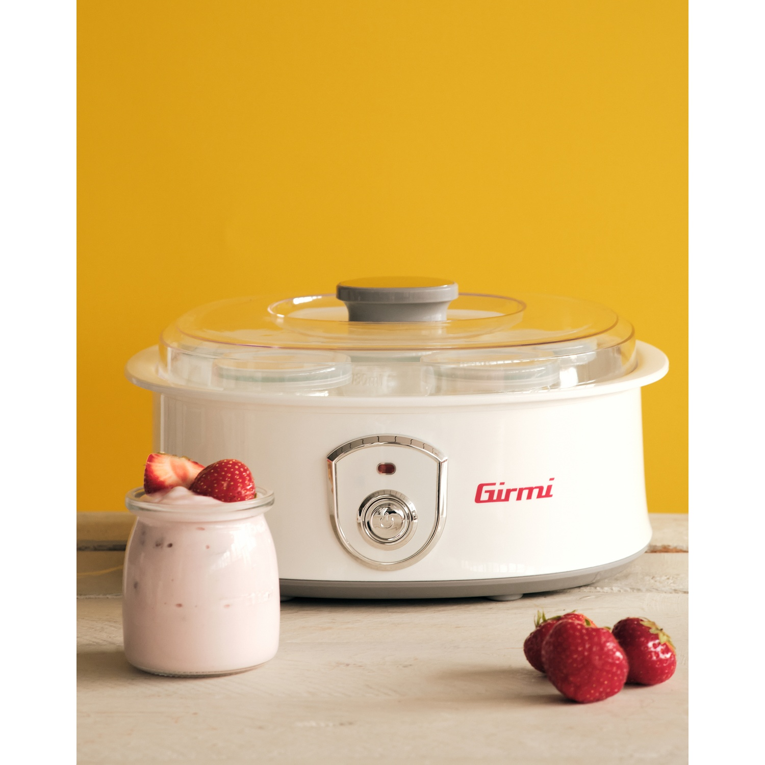 Una yogurtiera bianca con fragole e yogurt in un bicchiere, e fragole fresche, su una superficie di legno, con sfondo giallo.
