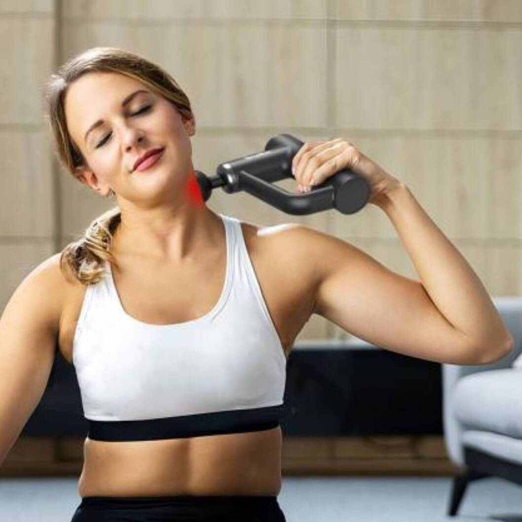 Eine Frau benutzt eine Massagepistole an ihrem Hals. Sie trägt einen weißen Sport-BH und schwarze Shorts. Der Hintergrund ist ein Raum mit Couch.