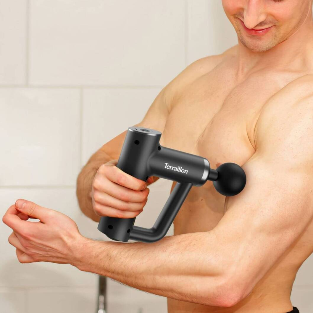Ein Mann benutzt eine schwarze Massagepistole an seinem Arm. Die Pistole ist mit 'Terraillon' beschriftet. Der Hintergrund ist eine weiße Kachelwand.