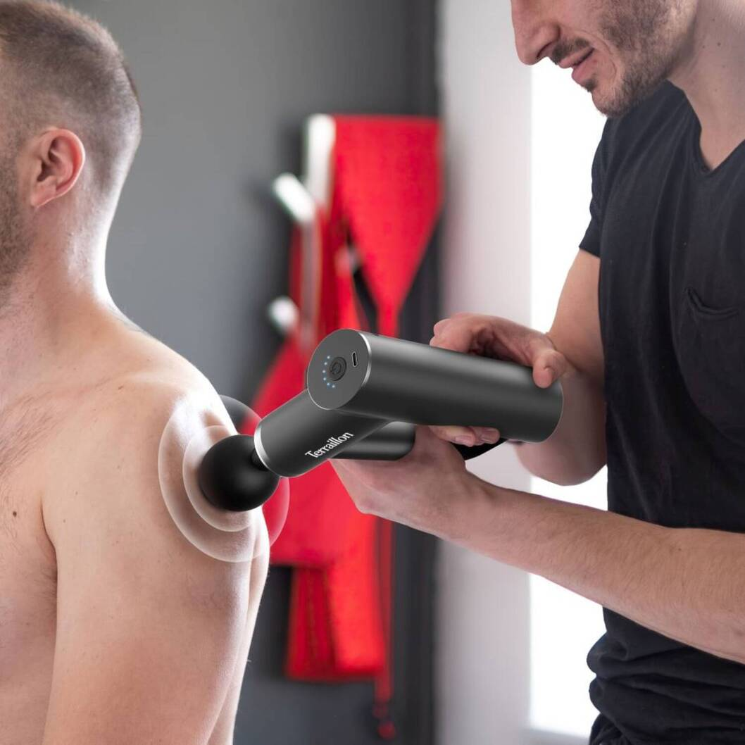 Ein Mann benutzt eine Massagepistole auf der Schulter eines anderen Mannes. Die Pistole ist schwarz, der Hintergrund grau mit roter Kleidung.