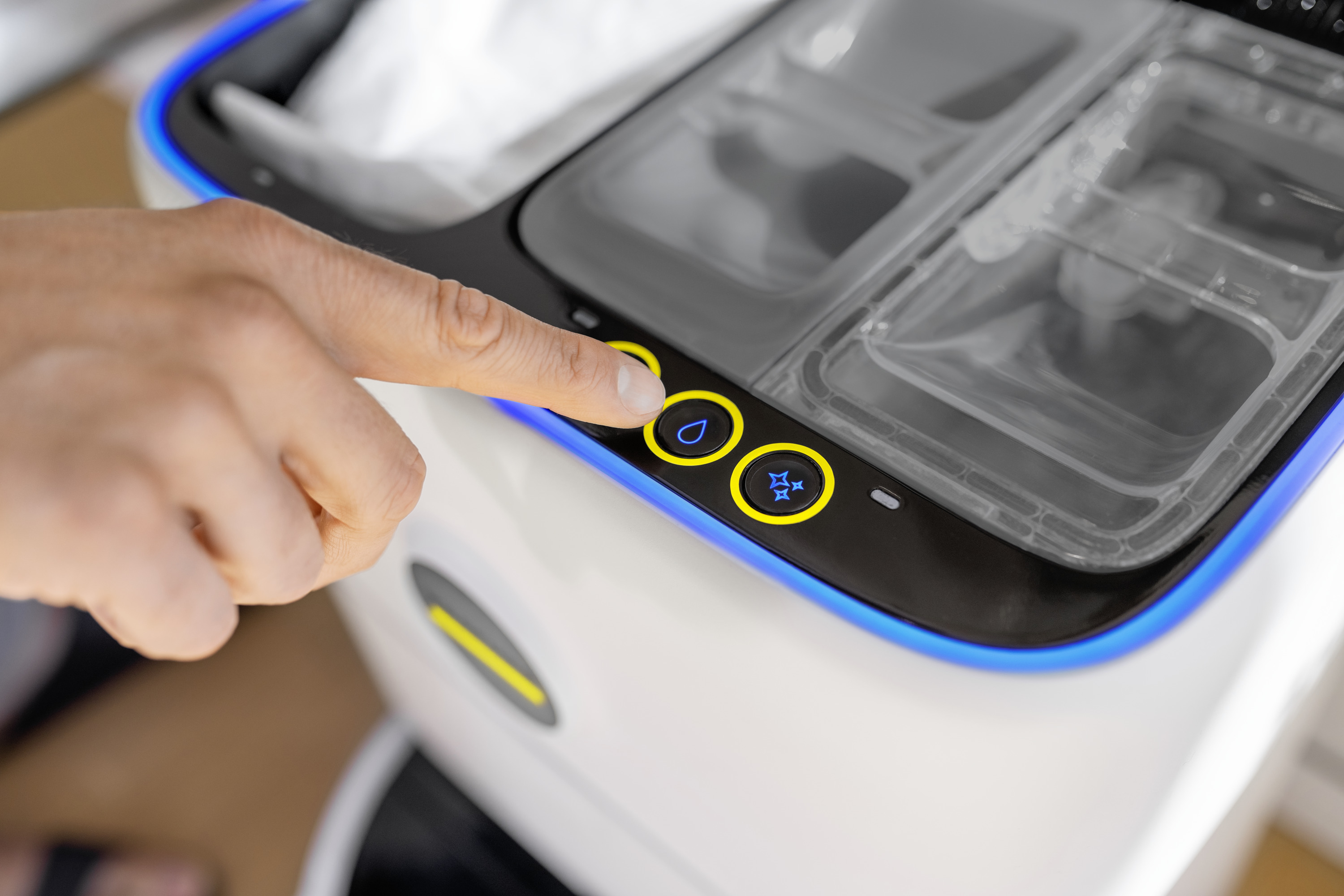 Palec naciska przycisk na białym urządzeniu. Przycisk ma ikonę kropli wody. Powyżej znajdują się pojemniki.